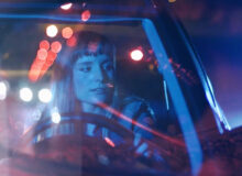 A lone woman drives anxiously. Lighting bokeh reflects in the windscreen.